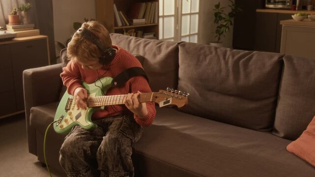 Cinematic shot of Caucasian young boy playing electric guitar at home and wearing headphones enjoying rhythm and music, copy space Useful for social media ads or vertical format campaigns