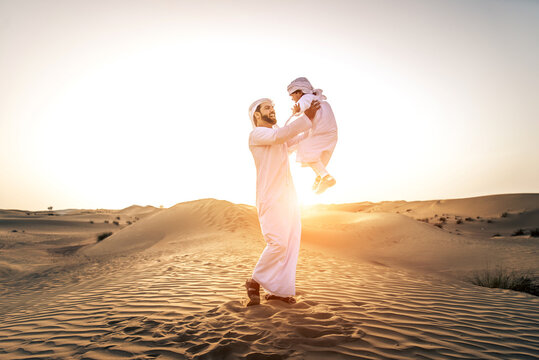 Arabian man and son playing in the desert at sunset - Middle-eastern father and his kid wearing traditional emirati kandura spending time together in Dubai desert