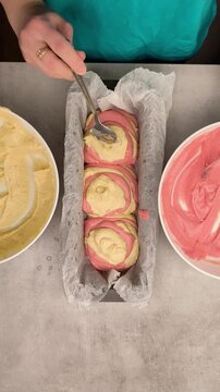 Top down view of a baker hands layering pink and yellow batter into a loaf pan lined with parchment paper, creating a beautiful pattern