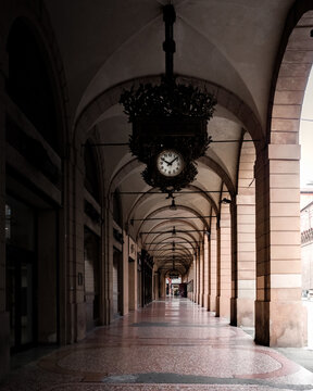 Bologna, Italy - 27 March 2022: View of a long, shadowed arcade, where the mosaic floor's intricate patterns lead the eye towards a distant, glowing exit beneath the antique clock.