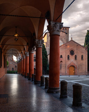 Bologna, Italy - 27 March 2022: View of the arched porticoes leading towards the Basilica di Santo Stefano, bathed in the warm glow of twilight against a textured sky.