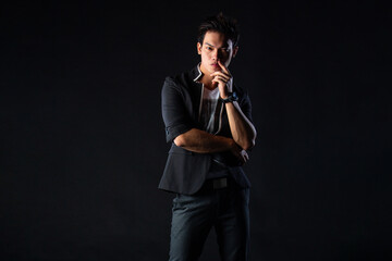 Fototapeta premium Young man poses confidently against dark studio backdrop, with dramatic lighting setup. Suit jacket layered over casual shirt, stylish yet edgy.