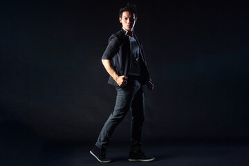 Fototapeta premium Young man poses confidently against dark studio backdrop, with dramatic lighting setup. Suit jacket layered over casual shirt, stylish yet edgy.