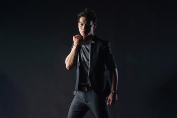 Obraz premium Young man poses confidently against dark studio backdrop, with dramatic lighting setup. Suit jacket layered over casual shirt, stylish yet edgy.