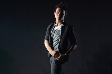 Fototapeta premium Young man poses confidently against dark studio backdrop, with dramatic lighting setup. Suit jacket layered over casual shirt, stylish yet edgy.