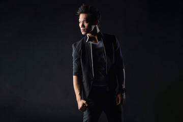 Fototapeta premium Young man poses confidently against dark studio backdrop, with dramatic lighting setup. Suit jacket layered over casual shirt, stylish yet edgy.