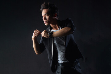 Fototapeta premium Young man poses confidently against dark studio backdrop, with dramatic lighting setup. Suit jacket layered over casual shirt, stylish yet edgy.