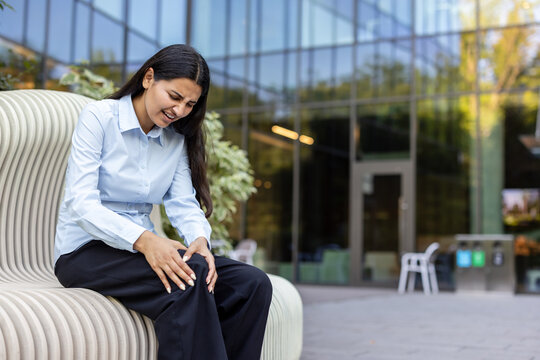 Young businesswoman sitting outside an office building, holding her knee with a pained expression, suffering from joint stiffness or injury while working in a hectic environment