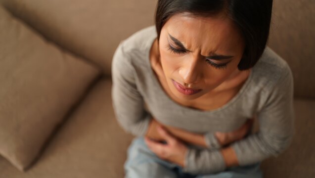 A woman experiencing stomach pain sits on a couch, clutching her abdomen in distress.
