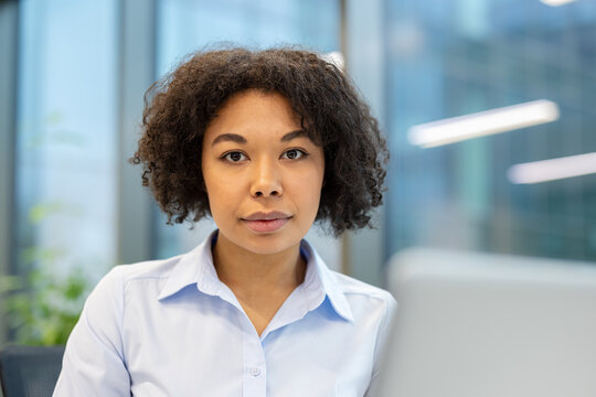 Close-up portrait of a young serious and focused African American woman sitting at a desk with a laptop and looking confidently at the camera