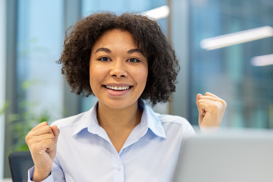 Close-up portrait of a happy young businesswoman in the office, sitting at a laptop and rejoicing in success, raising her arms up