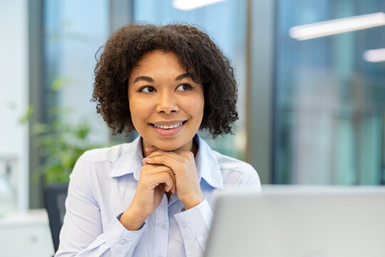 Smiling and thoughtful young African American woman sitting at laptop in office, holding hands together and looking aside
