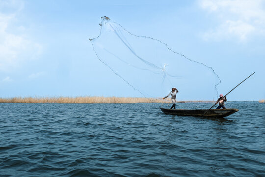 Ganvie, Benin - 14 April 2023: View of fishermen casting a net from a narrow boat on the shimmering waters, reeds providing a textured backdrop against the pale sky.