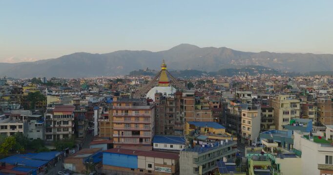 Aerial view of revealing of Boudhanath Stupa, Kathmandu Nepal, a buddhist monument with prayer flags rising through dense city buildings toward surrounding hills under warm golden hour evening sky.