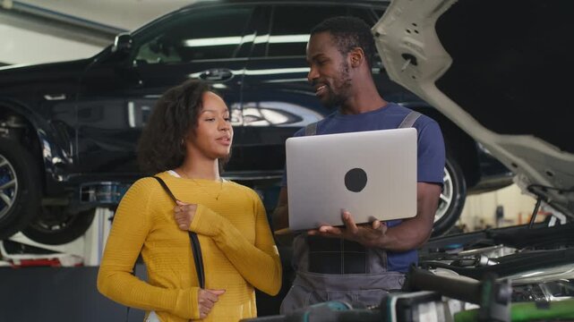 Professional mechanic in a workshop using a laptop for modern vehicle diagnostics, showing the woman client her car engine data.