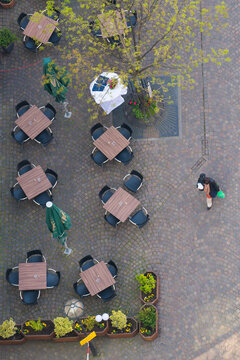 Frankfurt am Main, Germany - 10 April 2017: View of an outdoor cafe's brown tables and dark chairs on textured cobblestones, with lush green foliage and a solitary figure.