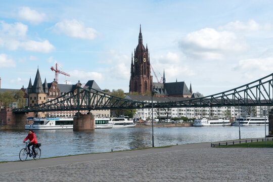 View of a cyclist pedals along the riverbank in Frankfurt, with the Eiserner Steg bridge and Frankfurt Cathedral rising against the sky, Frankfurt am Main, Hessen, Germany.