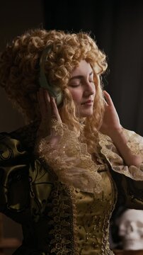 Vertical shot of relaxed young lady wearing curly wig and embroidered rococo dress enjoying music in wireless headphones