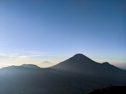 Dramatic silhouette of Mount Sindoro at sunrise with clear blue sky and misty valley from Sikunir hill viewpoint, Beautiful morning view of Mount Sindoro in Central Java under deep blue sky