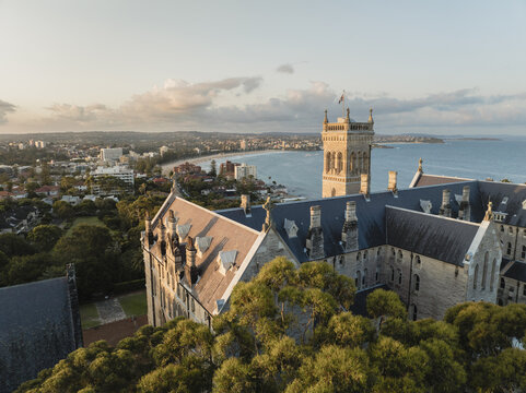 Aerial view of the iconic St Patrick's Seminary, its stone facade contrasting against the turquoise ocean, framed by lush greenery and distant coastline, Manly, New South Wales, Australia.