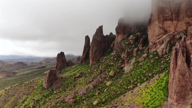 Aerial view of dramatic reddish-brown rocky peaks emerging from vibrant green slopes under a brooding, misty sky, Apache Junction, Arizona, United States.