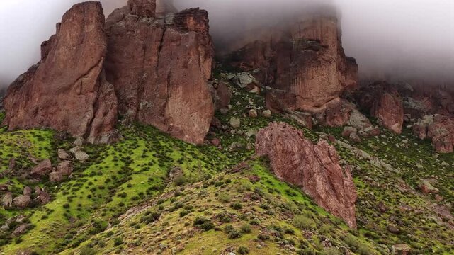 Aerial view of imposing reddish-brown rock formations emerging from a misty cloud, contrasting with vibrant green vegetation on the slopes, Apache Junction, Arizona, United States.