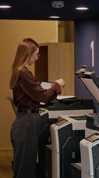 Vertical shot of young woman holding folder, walking through office and operating printer while copying business document