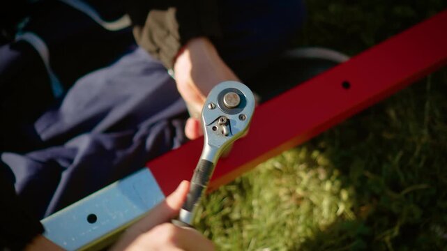 Manual worker tightening bolt with ratchet wrench on red metal beam in outdoor setting. DIY construction assembly repair project with mechanic tool equipment and craftsmanship detail in sunlight. Hand