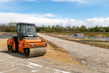 A steamroller stands still on a construction site next to a dirt road with trees and cloudy skies in the background © ProStockPhoto