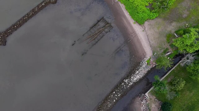 Aerial view of a tranquil, muddy shoreline with a rustic wooden structure, bordered by lush green foliage and a rocky barrier, Nyack, New York, United States.