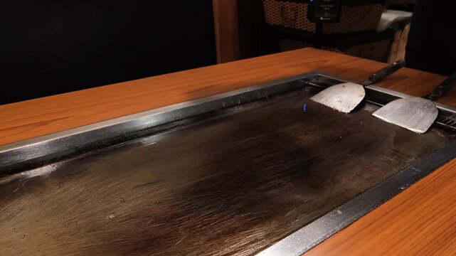 Close-up of a Japanese teppanyaki chef's hands using dual metal spatulas to scrape and prepare a large, seasoned iron flat-top grill for cooking in a restaurant.