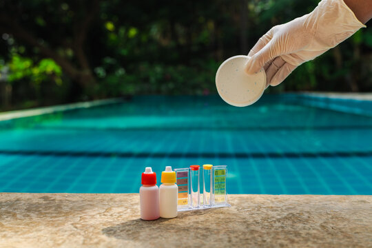 Gloved hand holding round chlorine tablet above swimming pool edge with water test kit and reagent bottles,