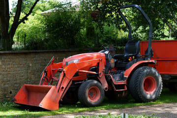 Tractor by wall. Garden vehicle beside stone barrier. Vintage utility tractor near landscaped yard. Classic garden tractor positioned on manicured grass with sunlight filtering through leafy trees. © Sasha Bond
