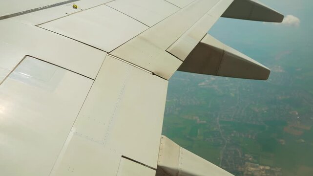 Airplane flying above city outskirts with wing and flap fairings visible. Aeroplane cruising over suburban area showing aerodynamic parts and control structures. Jet soaring through air displaying