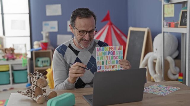Man holding colorful alphabet chart and pointing with pen at letters beside laptop in school building; teaching focus.