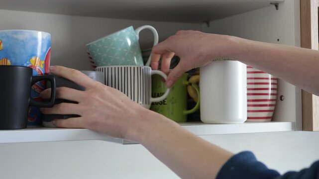 Hand reaching into cabinet to select a colorful mug from a shelf of kitchenware