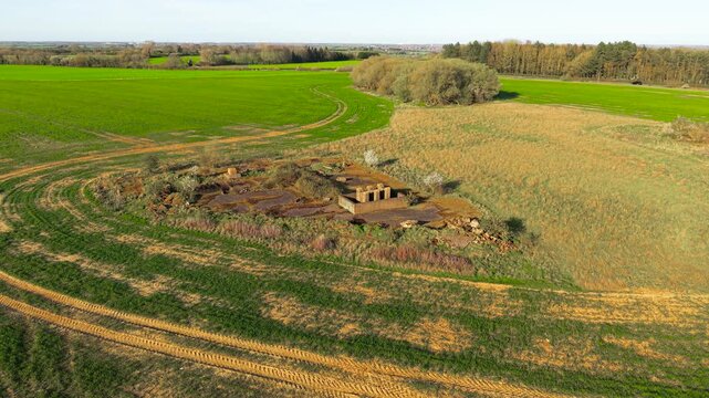 Abandoned military site with ruined bunker complex embedded in agricultural fields and winding tractor paths. Wide drone perspective revealing contrast between green crops and decaying structures