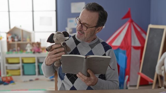 Man reading book holds elephant puppet in school building reading aloud at a table in colorful kindergarten playroom; warmth storytelling.