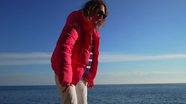 Woman beach jacket standing on a sunny coastline wearing a vibrant pink puffer coat and looking down near the ocean