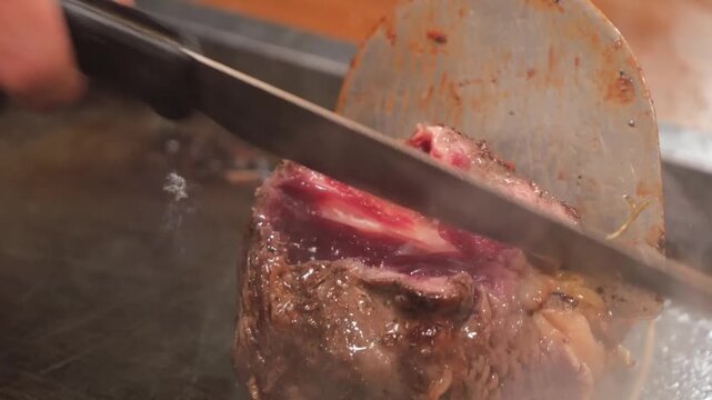A professional chef uses a metal spatula to flip and sear a thick, juicy cut of premium Wagyu beef on a steaming hot teppanyaki grill. The footage captures the sizzling texture and rich marbling of th