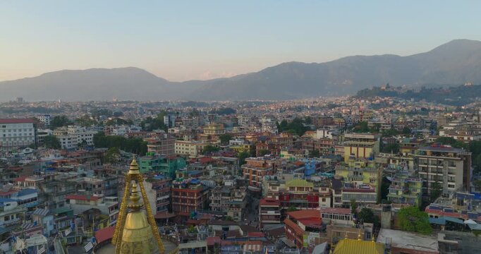 Aerial view of revealing the golden top of Boudhanath Stupa, Nepal rising above dense city buildings with prayer flags, birds and distant hills under a soft sunset sky.