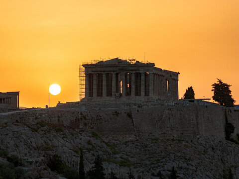 View of the Parthenon's ancient silhouette against a vibrant golden sunset sky above the rugged Acropolis, Athens, Central Athens Regional Unit, Greece.
