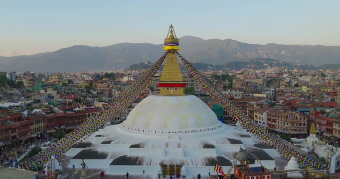 Aerial view of the white Buddhist monument and golden spire of Boudhanath Stupa at the dense city with long prayer flags stretching toward surrounding hills under soft evening sky in Kathmandu Nepal.