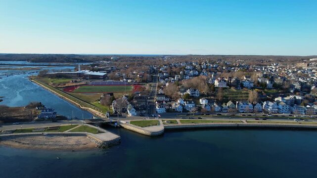 Drone view of Gloucester Massachusetts town with sunny waterfront, blue harbor water, neighborhoods and scenic coastline creating classic New England coastal city atmosphere on clear day. Lateral wide