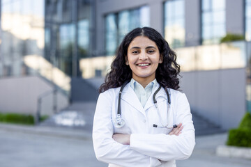 Young indian female doctor in a white lab coat standing with arms crossed, smiling confidentially...