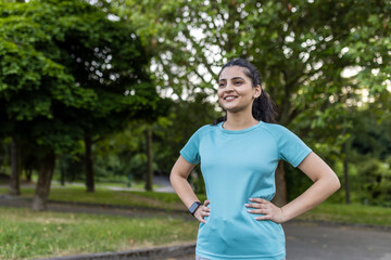 Obraz premium Young indian woman in sportswear stands with hands on hips, smiling and looking away in a green urban park, enjoying a confident, healthy, active lifestyle and fresh air