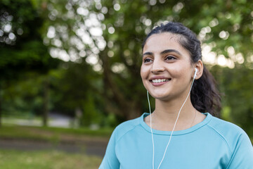 Naklejka premium Young indian woman smiling cheerfully outdoors, listening to music with earphones, enjoying fitness and a healthy lifestyle in a green park environment