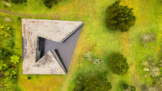 Aerial view of a geometric concrete building with a dark, reflective opening, amidst vibrant green and earthy fields, Ponta Delgada, Azores, Portugal.
