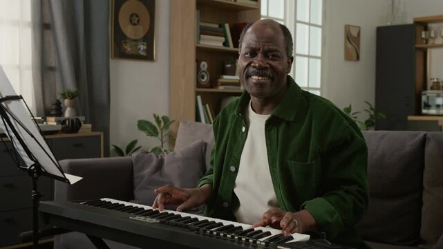 Medium shot of excited senior African American man learning to play keyboard at home and turning looking at camera with smile, reading notes Ideal for active seniors retirement programs