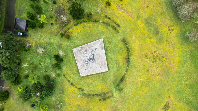 Aerial view of a weathered square pyramid-like structure encircled by ancient stone rings, set against a vibrant tapestry of green and yellow fields, Ponta Delgada, Azores, Portugal.